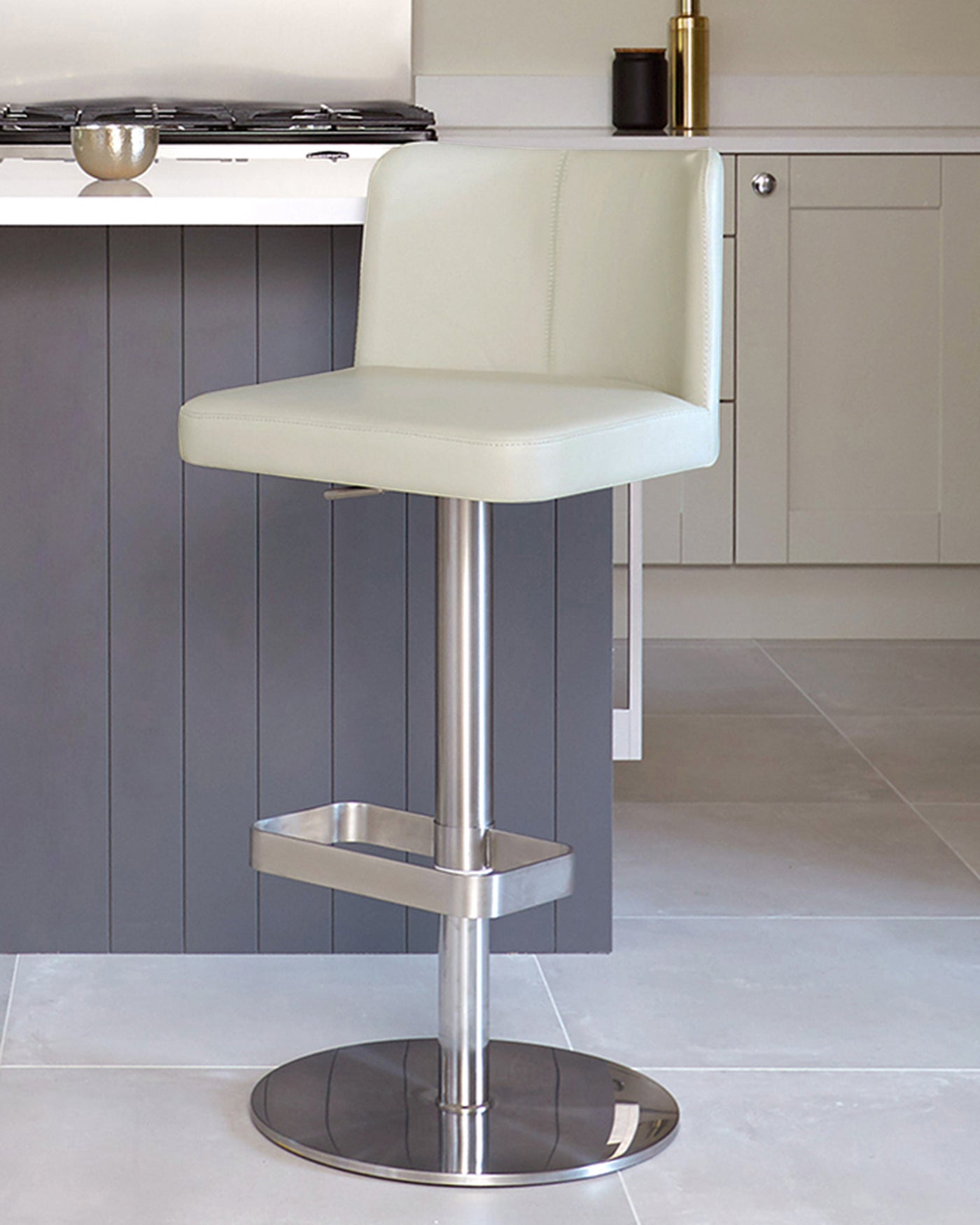 Modern bar stool with a cream faux leather seat, low backrest, and visible stitching details. The stool features a sleek chrome base, pedestal, and footrest. The design is contemporary and suitable for kitchen or bar counters.
