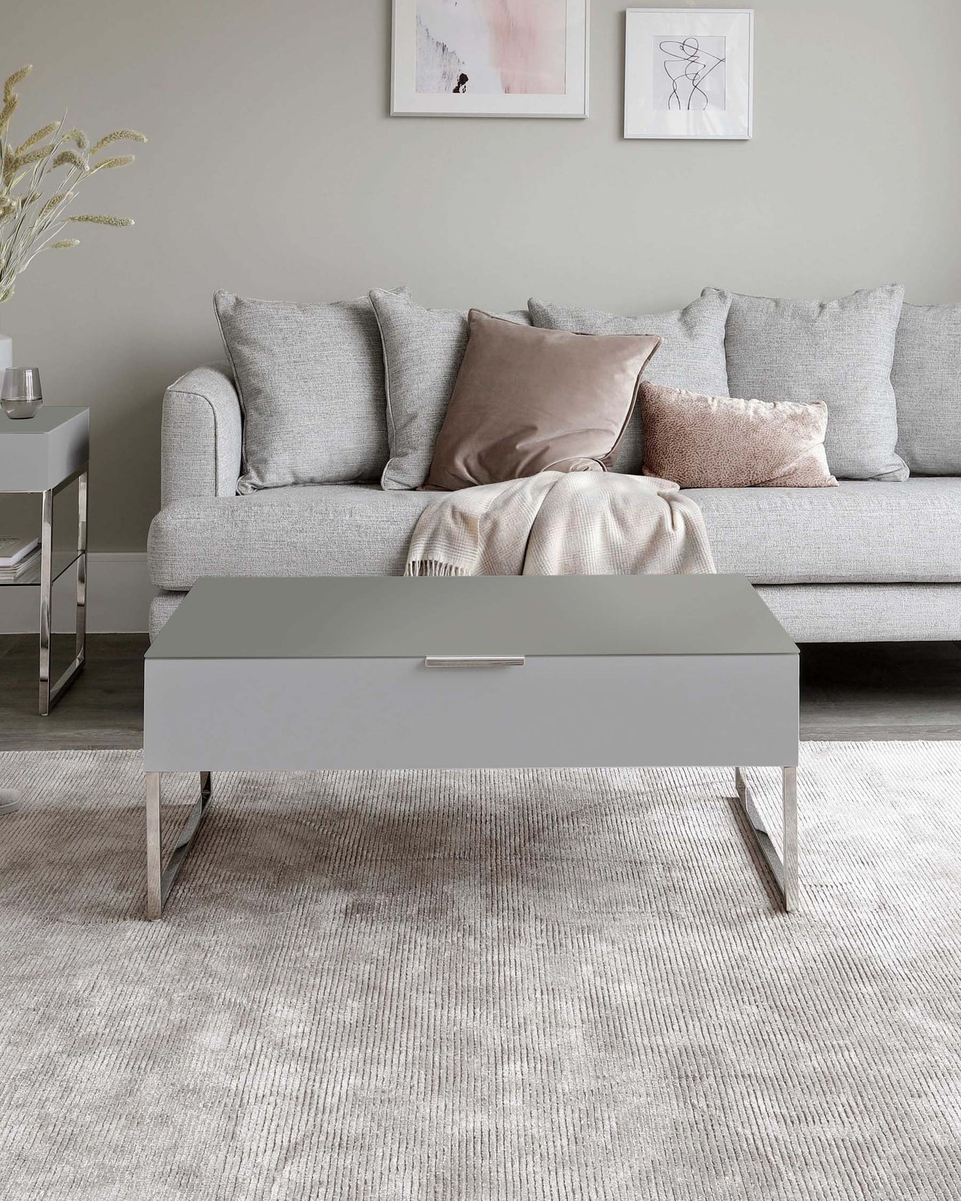 Contemporary grey fabric sectional sofa adorned with a variety of pillows in muted tones and a light throw blanket. In the foreground, a sleek grey coffee table with a minimalist design and metal legs sits atop a textured off-white area rug. A small modern side table in grey with a reflective glass tabletop is partially visible beside the couch.