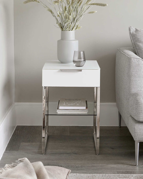 Modern white lacquered side table with chrome legs and an open lower shelf, displayed alongside a grey fabric upholstered armchair. The table is accessorized with a large matte grey vase holding dried foliage and a simple clear glass.