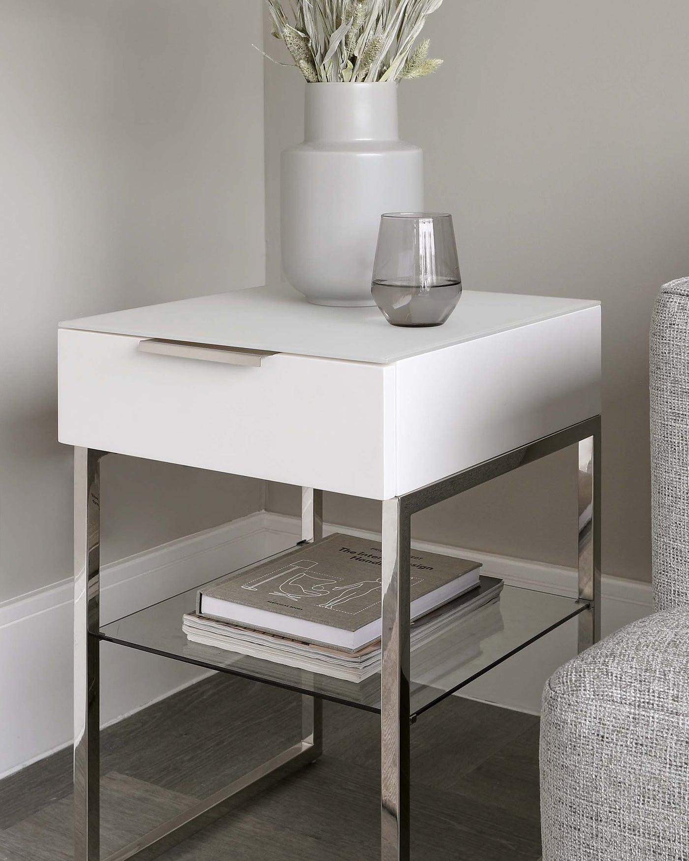 Modern bedside table with a white, square top drawer, sleek silver handle, and stainless steel frame. Features a lower glass shelf suitable for books or decorative items. Positioned next to an upholstered chair, with decorative vase and glass atop the table.