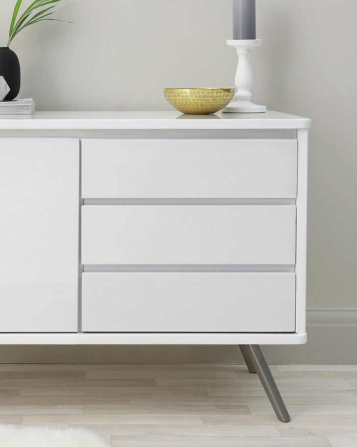 Modern white dresser with clean lines, featuring three drawers with no visible handles, angled metal legs, and a smooth finish. Accessorized with a decorative bowl and candle holder on top.