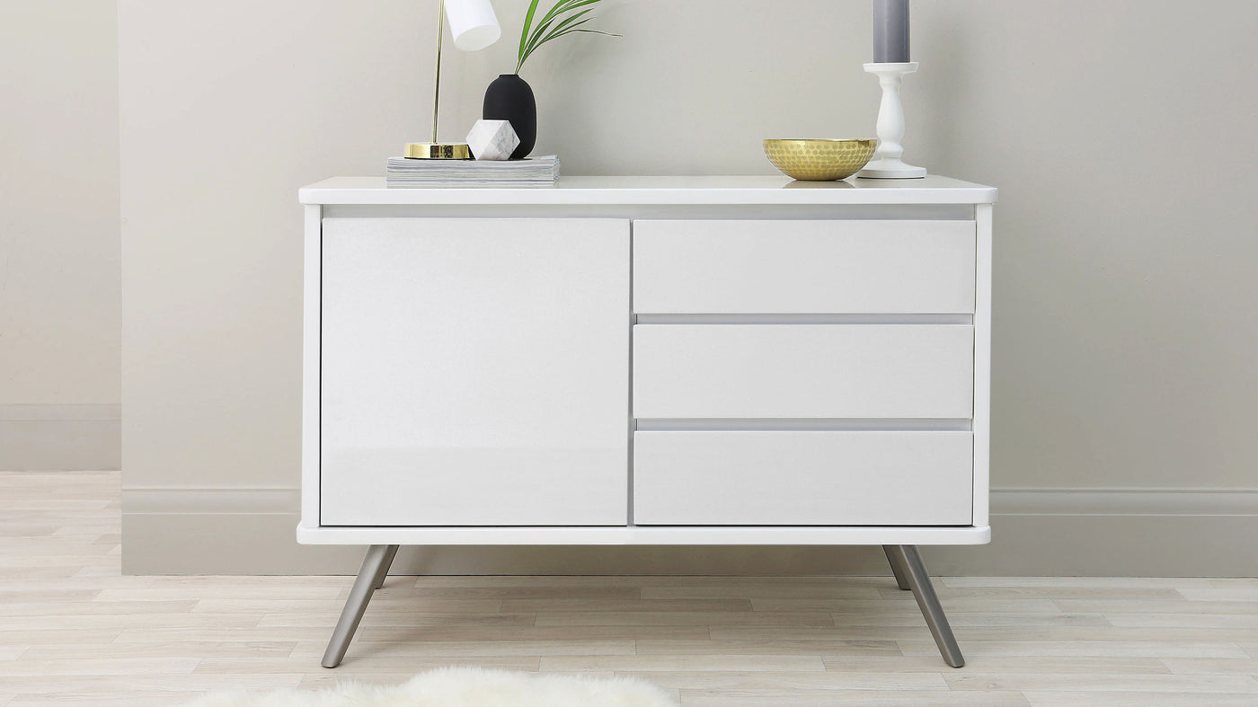 White modern minimalist sideboard cabinet with two doors and three drawers on angled metal legs in a light, neutral interior setting. A lamp, vase with greenery, stack of books, and decorative bowl adorn the top.