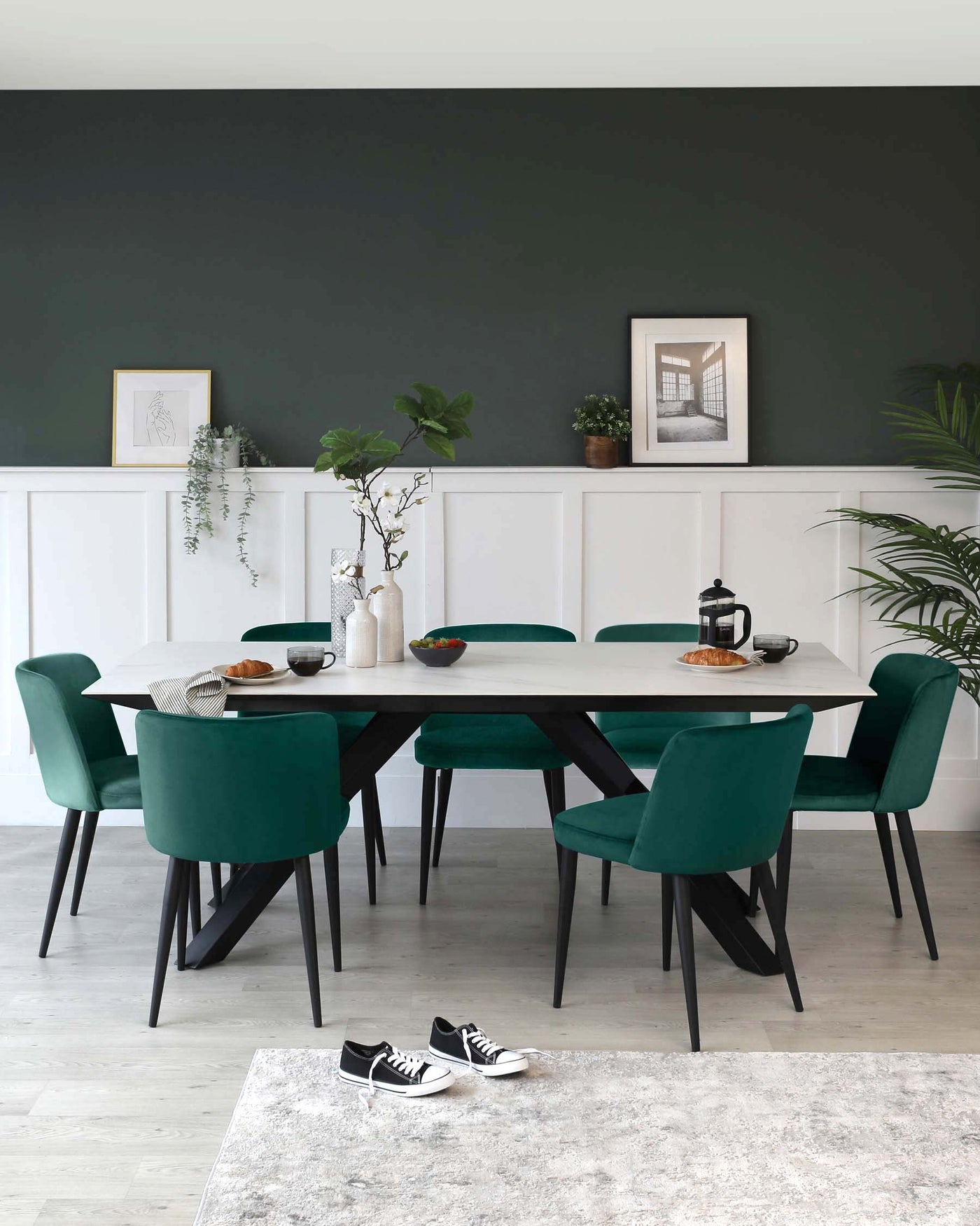 Contemporary dining room featuring a rectangular white tabletop with black legs, surrounded by six plush teal upholstered chairs with black wooden legs. A minimalistic rug with light grey tones anchors the dining set on a light wooden floor.