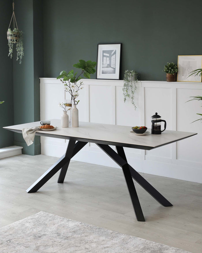 Modern minimalist dining table with a rectangular light grey tabletop and distinctive geometric black metal legs, set in an elegantly styled room with white and green decor.