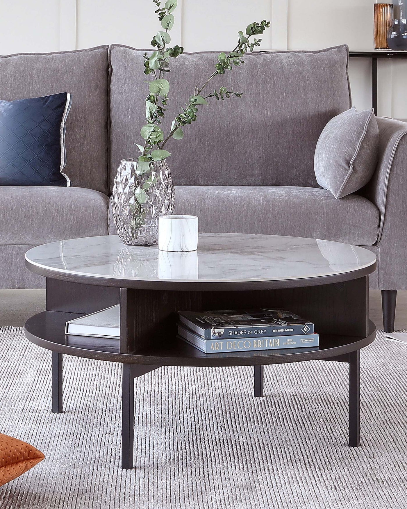 A modern living room showcasing a sleek, round, dark wooden coffee table with a glossy white marble tabletop. Below the tabletop is a circular shelf holding books, emphasizing the table's dual-function design. Adjacent to the table is a section of a grey fabric sofa with a plush cushion and a decorative pillow. The setting is completed with a textured light grey area rug, providing a soft foundation for the elegant furniture pieces.