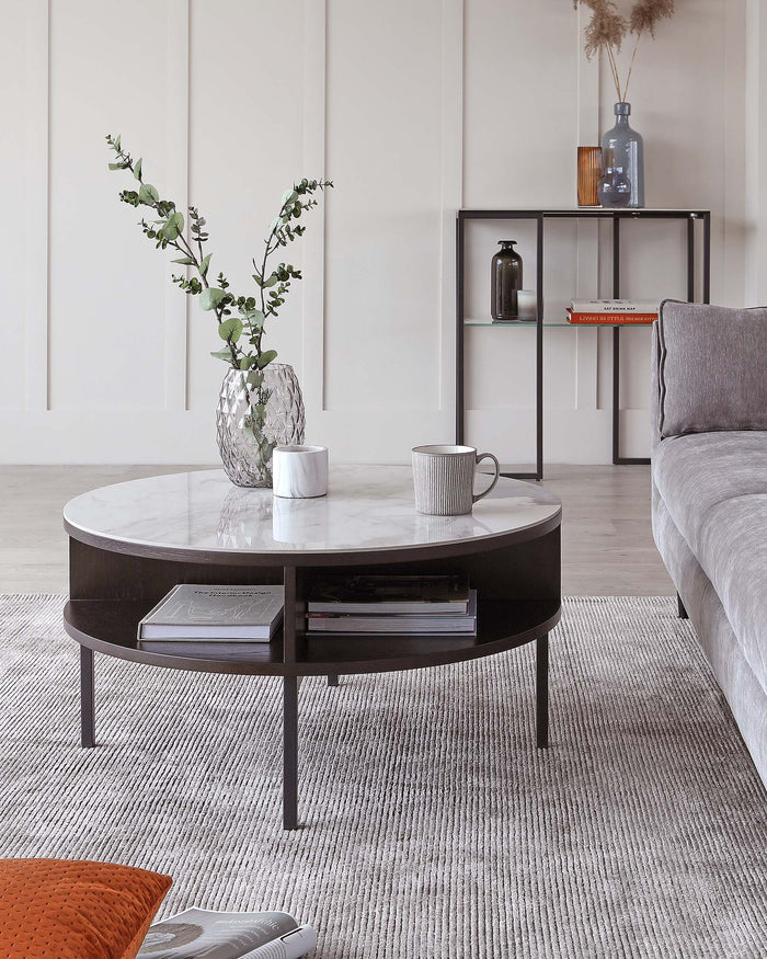 A modern living room featuring a round marble-top coffee table with a dark wooden base and a lower shelf. Beside it, a minimalistic, metal frame console table with glass shelves displays decorative items. A corner of a grey upholstered couch is partially visible to the right. The furniture pieces are on a textured grey area rug.