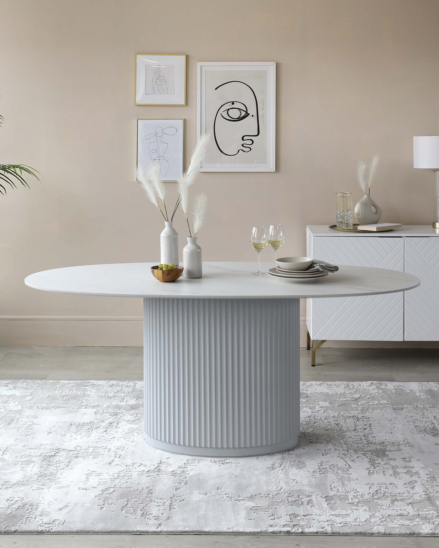 Modern minimalist dining room featuring an oval white table with a fluted pedestal base, accompanied by a sleek white sideboard with subtle geometric patterns. The room is accessorized with decorative vases and a tasteful arrangement of wall art.