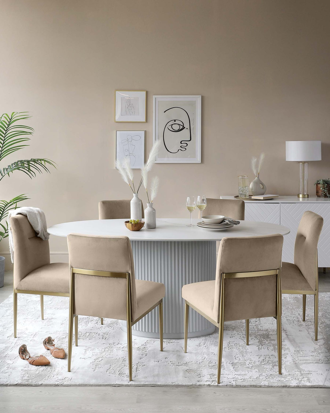 Elegant dining room furniture set featuring a modern round table with a fluted pattern on its pedestal base, finished in white. Accompanied by four plush, taupe upholstered chairs with sleek golden metal legs, positioned on a textured white area rug.