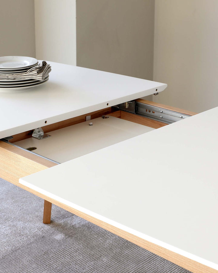 Modern extendable dining table with a white tabletop partially extended to reveal the mechanism underneath, showing metal slides and wooden support structure. The table rests on light wooden legs and is set against a neutral background, with a stack of white plates and a cloth napkin on the surface indicating its use in a dining setting.