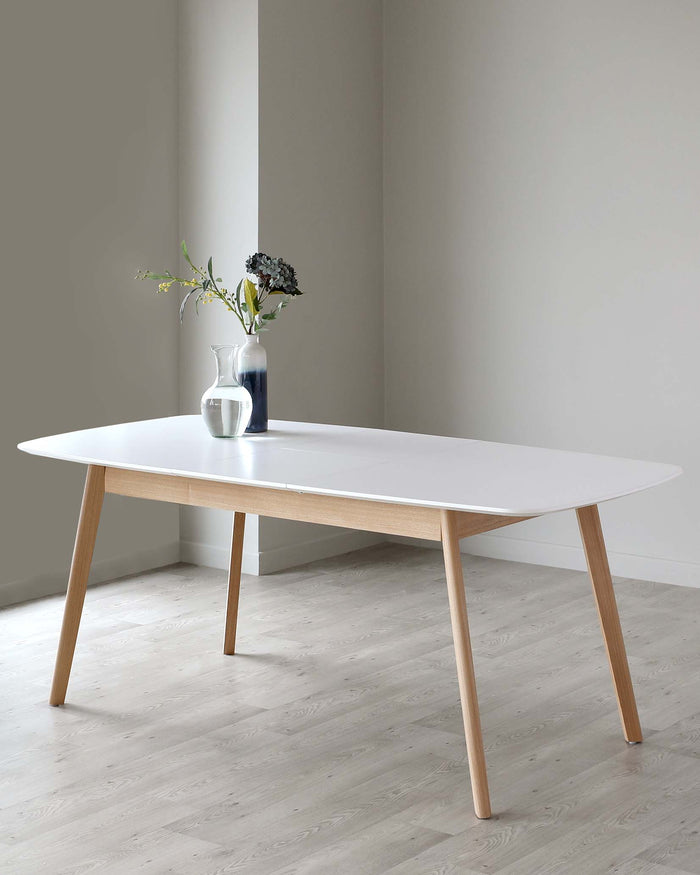Modern minimalist white oval dining table with natural wood tapered legs in a clean, neutral interior setting. A vase with flowers adds a touch of decor.