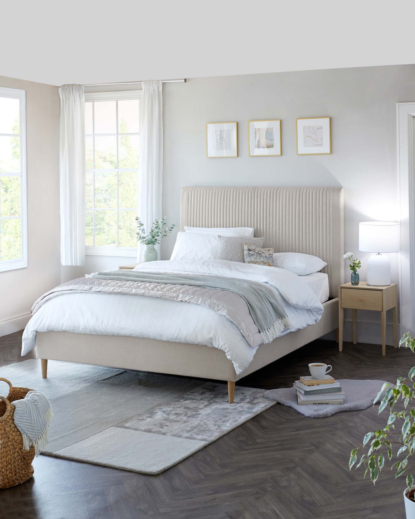 Elegant contemporary bedroom featuring a beige upholstered platform bed with a vertically channelled headboard, complemented by a pair of natural-finished wooden bedside tables with a single drawer. A matching white ceramic table lamp is placed atop one of the tables, enhancing the room's modern aesthetic.