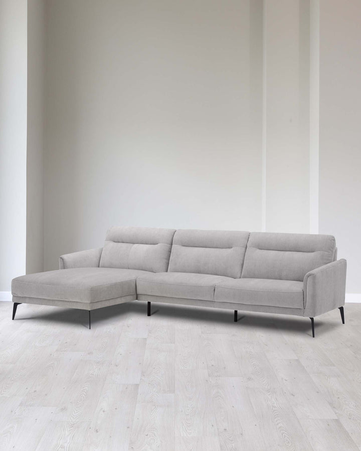 A modern light grey fabric sectional sofa with clean lines, featuring a chaise lounge on the left side, simple tufted backrest cushions, and minimalist black metal legs. The couch is set in a contemporary room with white walls and light wooden flooring.