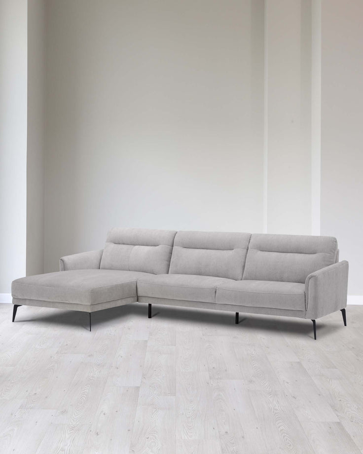 A modern light grey fabric sectional sofa with clean lines, featuring a chaise lounge on the left side, simple tufted backrest cushions, and minimalist black metal legs. The couch is set in a contemporary room with white walls and light wooden flooring.