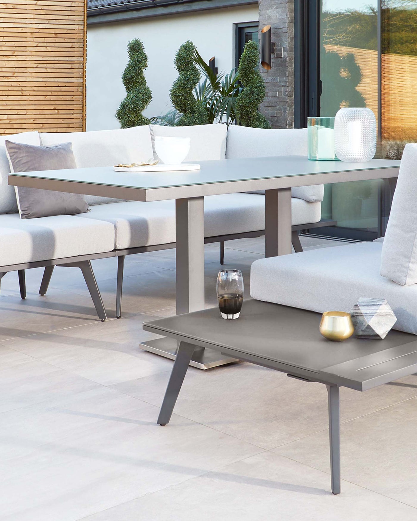 Outdoor modern furniture set featuring a sleek grey rectangular dining table and a matching corner sectional sofa with light grey cushions and plush back pillows. Accessories include a bowl, books, and decorative vases on the table, with a glass candle holder on the sofa-side table. The setup is complemented by topiary plants and a contemporary home facade in the background.