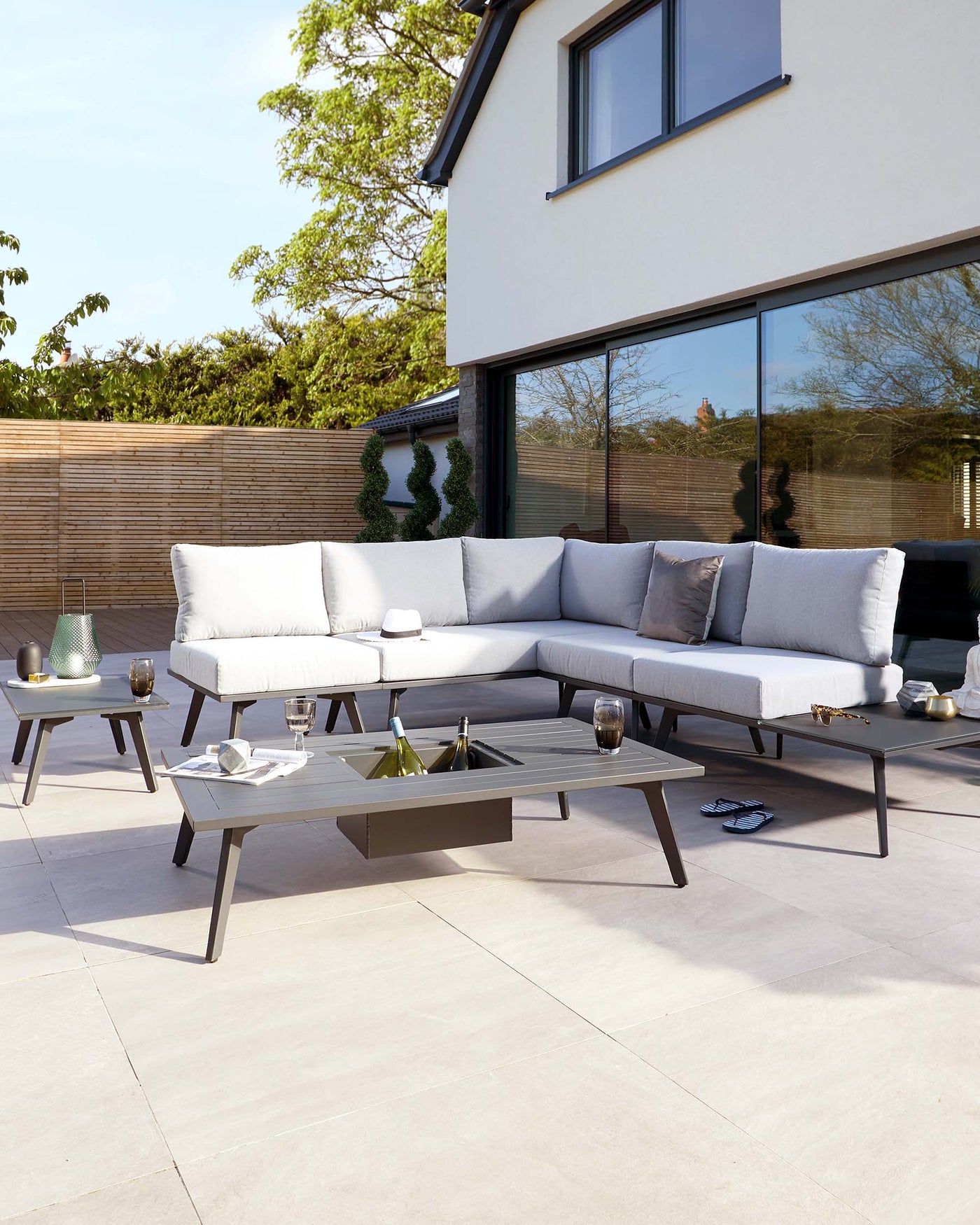 Modern outdoor furniture set featuring a sleek, taupe-coloured sectional sofa with plush light grey cushions and matching accent pillows, accompanied by two minimalist geometric side tables and a rectangular coffee table, all in a coordinating dark metal finish, displayed on an elegant patio.