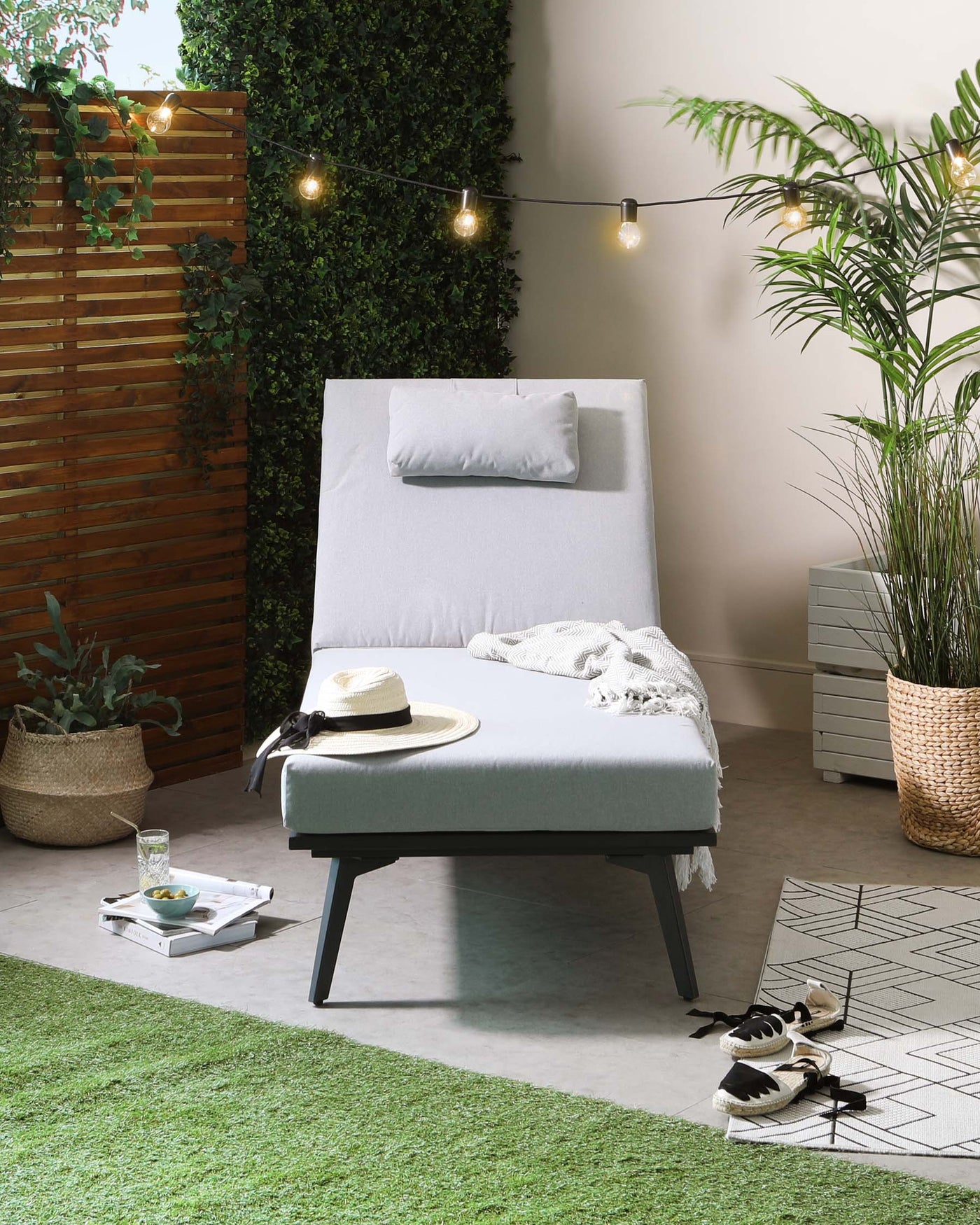 Modern outdoor chaise lounge with a light grey cushion and simple pillow on a sleek, black metal frame, accompanied by a small coordinating side table. The setting is complemented by decorative string lights, potted plants, a woven basket, and casual décor items for a relaxed patio ambiance.