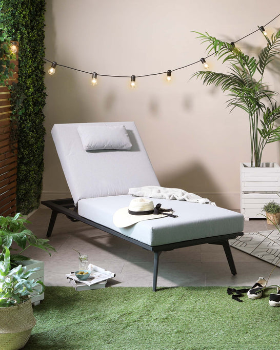 Modern outdoor chaise lounge with a sleek black metal frame and light grey cushioning, featuring an adjustable backrest, accompanied by a small white side table with a slatted design. The setting is enhanced with ambient string lights, potted plants, and decorative items for a cosy patio atmosphere.