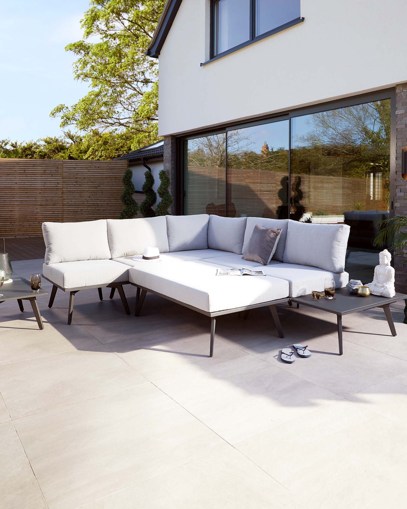 Modern outdoor L-shaped sectional sofa with light grey cushions on a minimalist dark frame, accompanied by a rectangular coffee table with the same design aesthetic. Two small side tables in a matching style flank the sofa, all displayed on a patio setting with decorative items and outdoor pillows completing the comfortable, stylish arrangement.