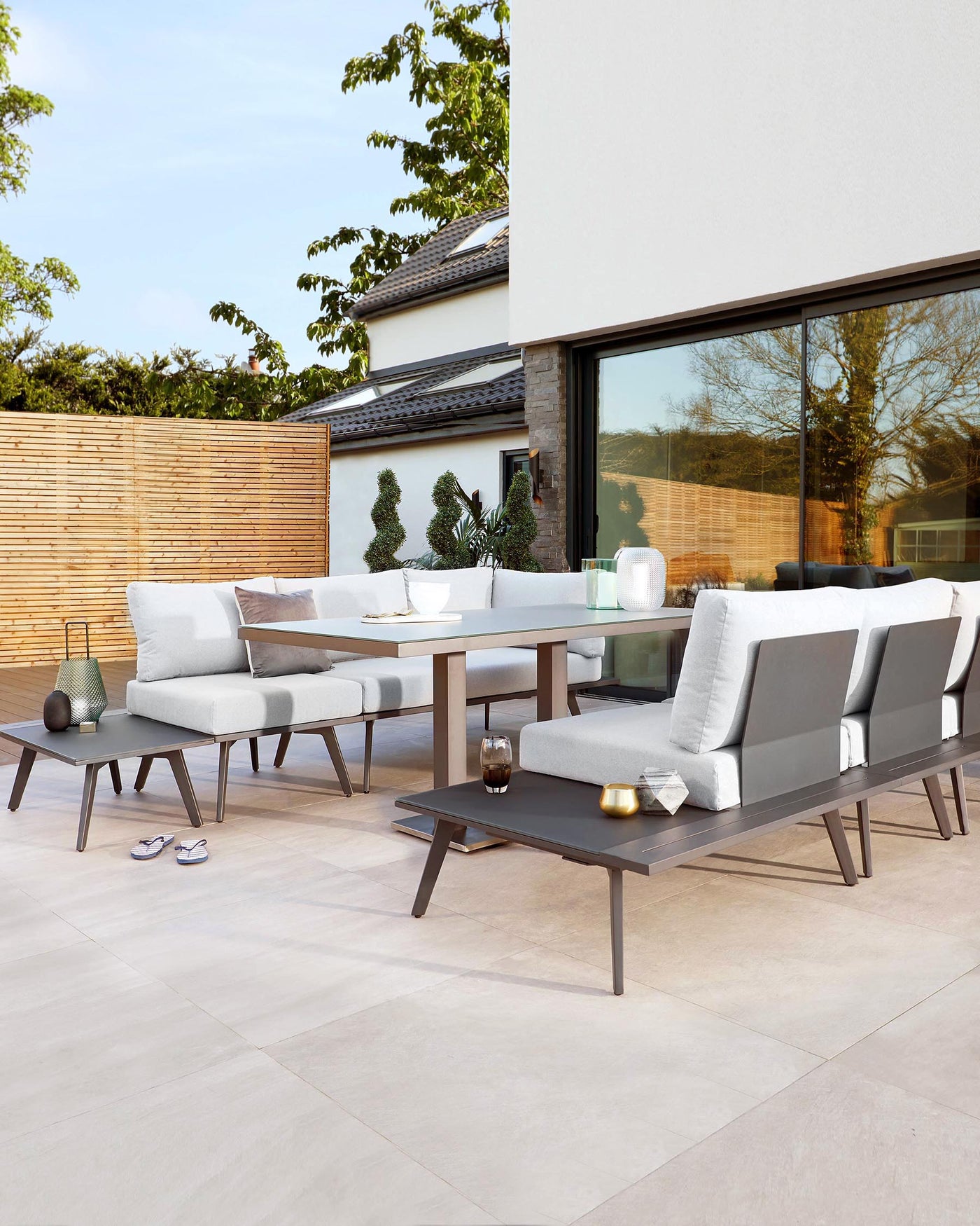 Modern outdoor patio furniture set featuring a rectangular dining table with a dark wood finish, accompanied by six contemporary style chairs with grey cushioned seats and backs. Two matching benches provide additional seating. A low-profile coffee table coordinates with the set, all arranged on a light tiled patio. Decorative items, including candles and a small vase, adorn the tables for a cosy ambiance.