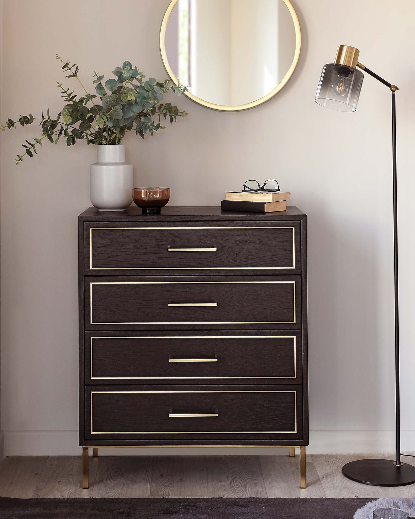 A contemporary dark wood five-drawer chest with brass-finished metal handles and legs, styled with a white vase of eucalyptus, a wooden bowl, books, glasses, and an adjacent floor lamp with a brass base and a clear shade. There is a circular gold-coloured framed mirror on the wall above the chest.