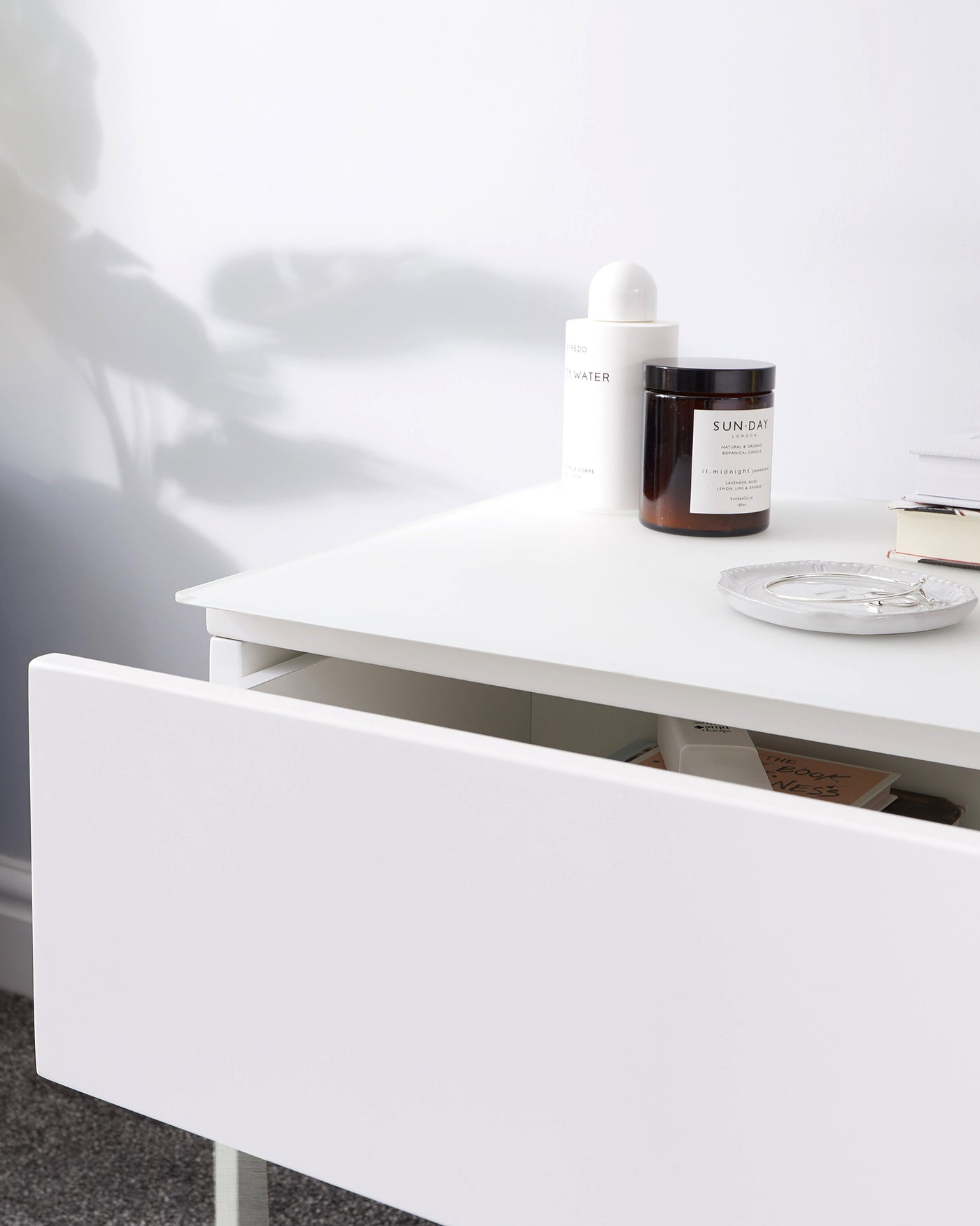 Modern minimalist white console table with clean lines and a smooth finish, featuring an open shelf with a partially opened drawer showing books and items inside. The tabletop is accessorized with a white cylindrical container, a brown glass jar, and stacked books next to a small decorative plate.