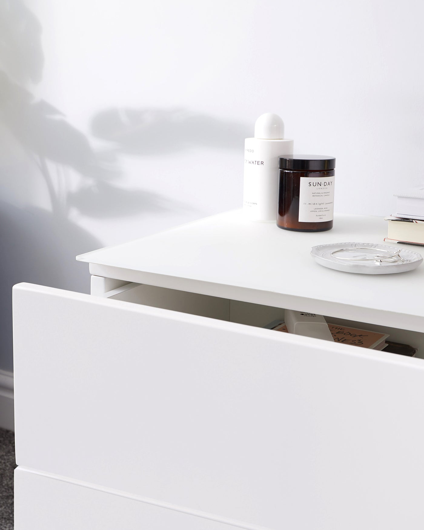 A modern white minimalist dresser with clean lines featuring flat-panel drawers that are partially open, showing stored items like books and miscellaneous objects. On top of the dresser sits two cosmetic bottles and a small ornamental dish. The simple design emphasizes functionality and contemporary elegance.