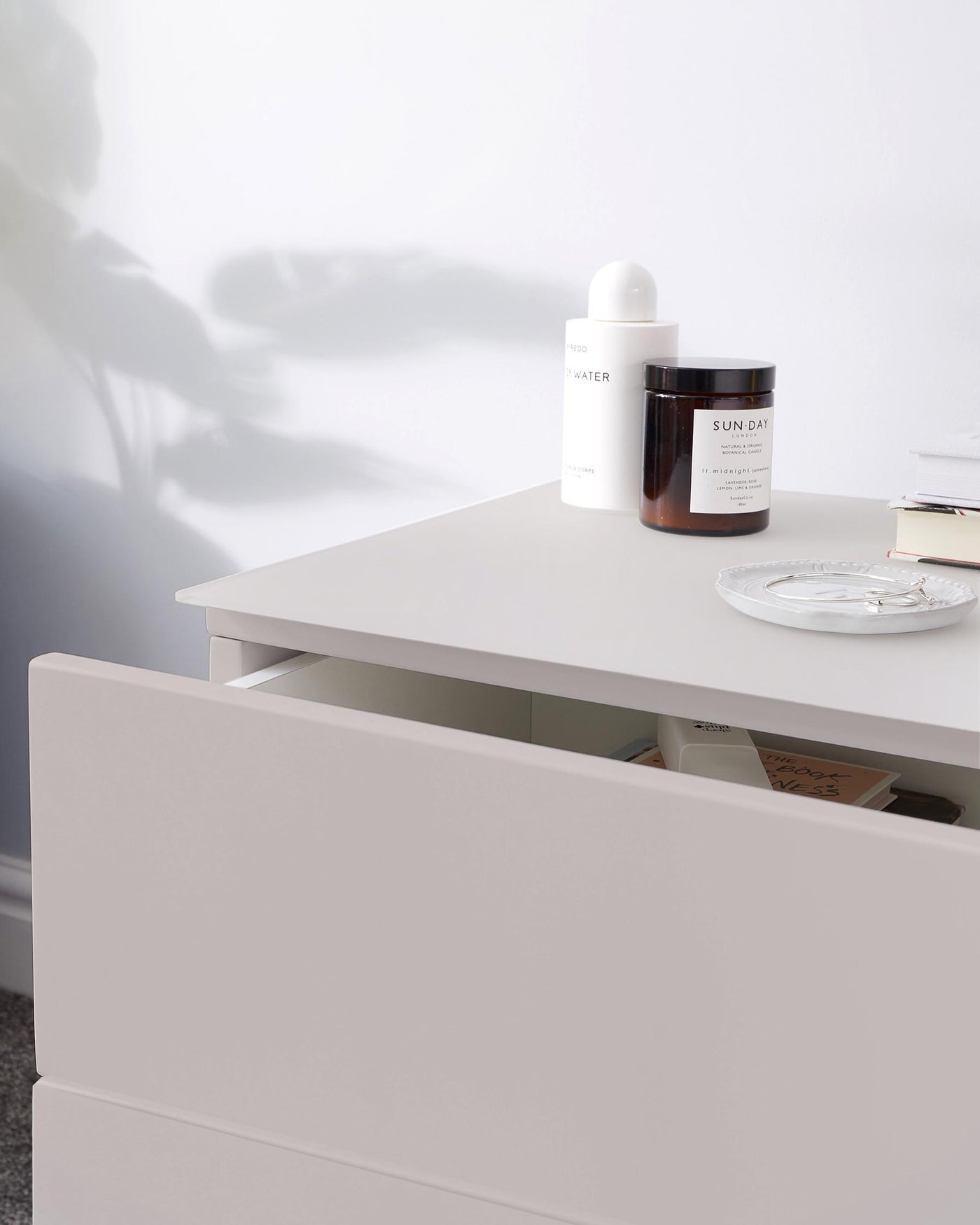 Modern minimalist white side table with a drawer partially open revealing books inside, along with a shelf holding various decorative items on the tabletop.