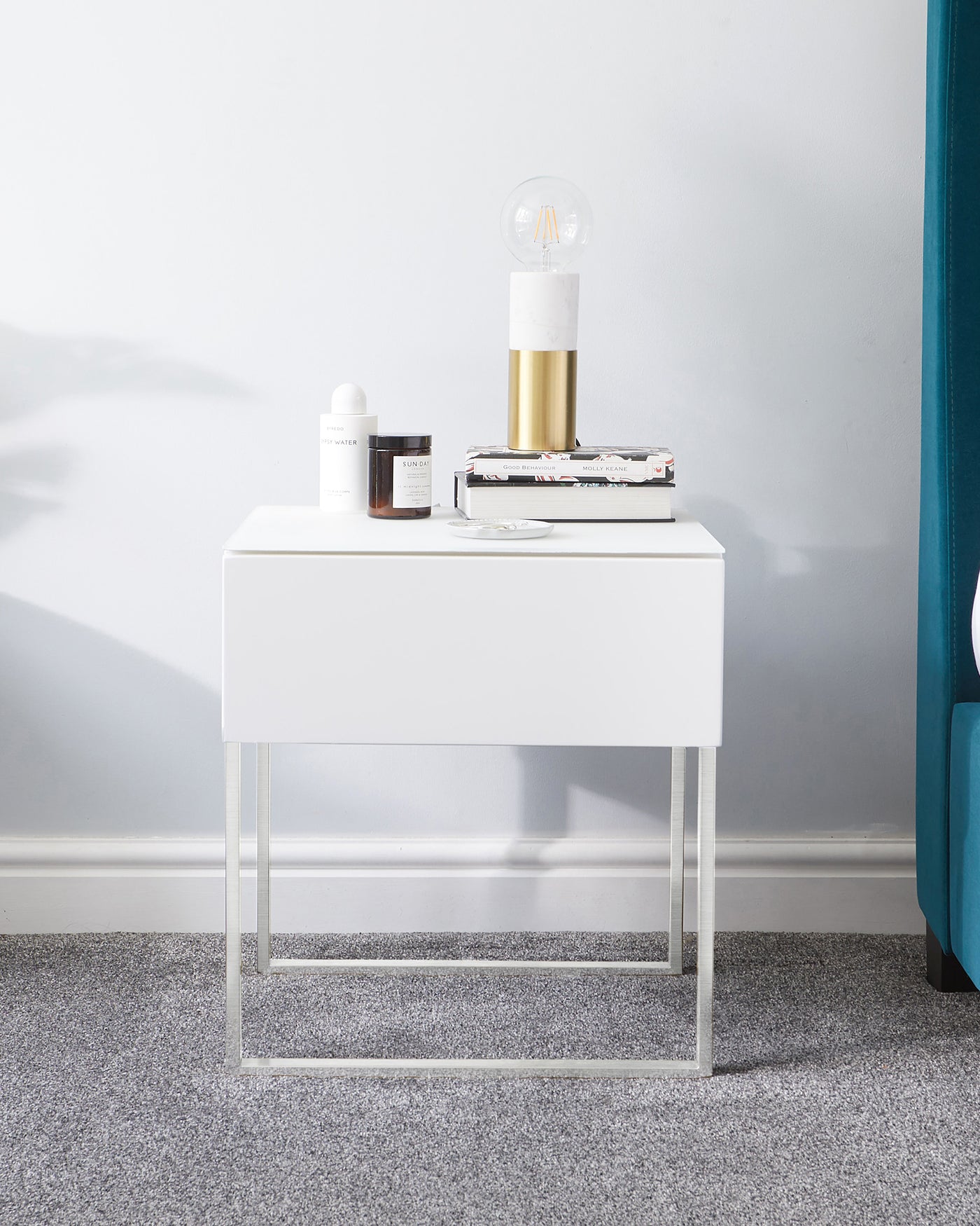 White modern minimalist nightstand with a single drawer, featuring a sleek design with a chrome-finished metal base. The tabletop is adorned with a small collection of items, including a lamp with a marble and gold base, as well as a stack of books and decorative jars.