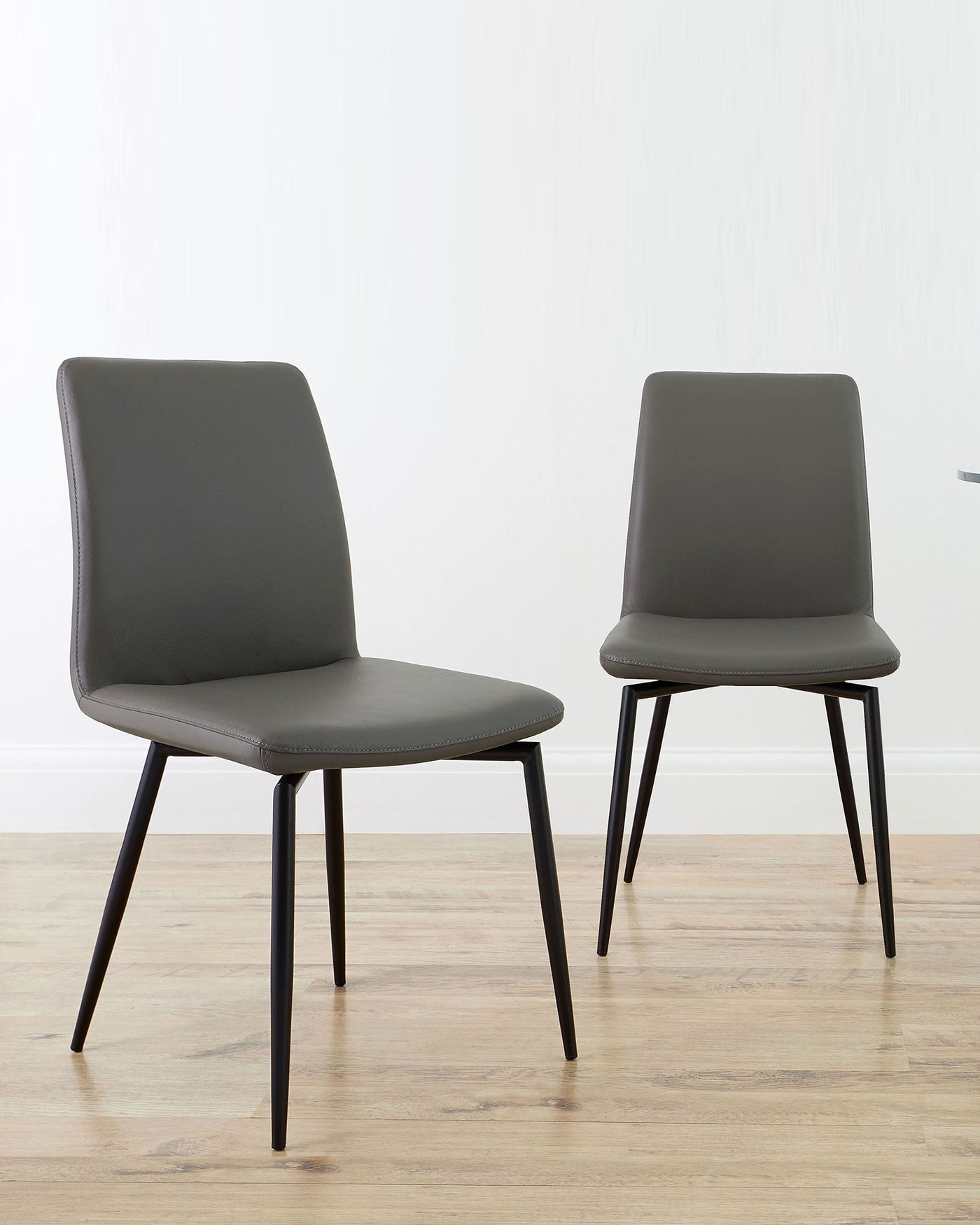 Two modern minimalist dining chairs with dark grey upholstery and black metal legs, positioned on light wood flooring against a white wall.