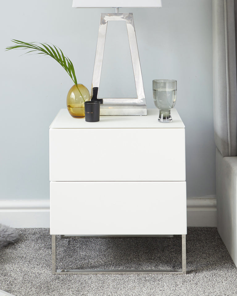 Modern white two-drawer nightstand with a sleek, minimalist design and metallic silver base frame, displayed in a contemporary bedroom setting.