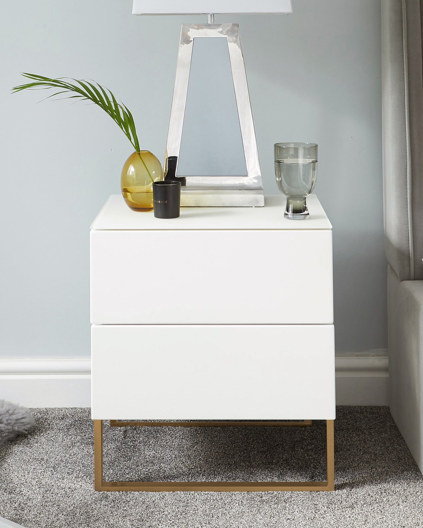 Modern white two-drawer nightstand with brass-finished metal base, styled with a decorative lamp, glass candle holder, and a vase with green foliage on top. The nightstand is set against a grey wall and complemented by a light grey carpet.
