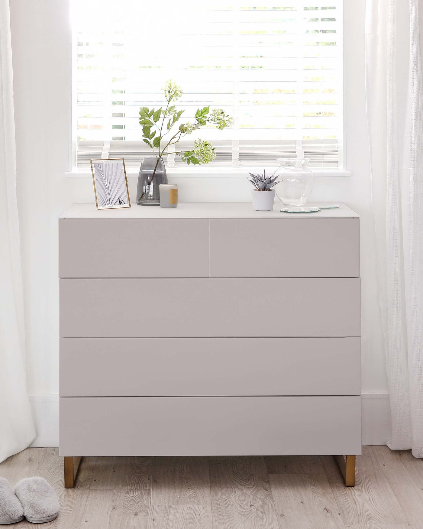 Modern light grey four-drawer dresser with angular lines, standing on four tapered wooden legs. The dresser is styled with a clear glass vase holding greenery, a small potted plant, a picture frame, and decorative items on its surface, against a backdrop of a bright window with white Venetian blinds and sheer curtains.