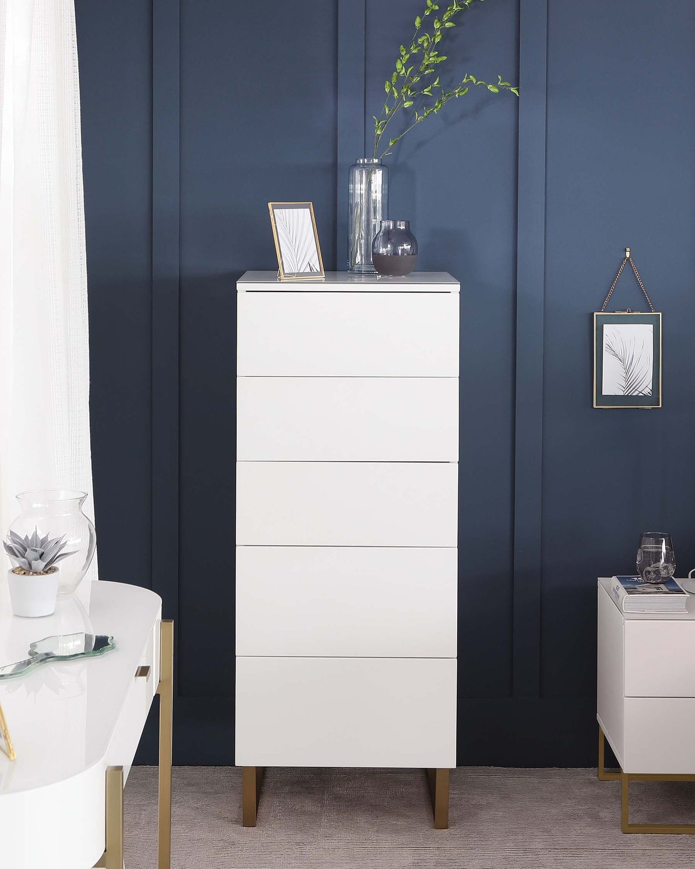 A modern white tallboy dresser with five drawers and gold-tipped legs against a dark blue wall. Decorated with a frame, a clear vase with greenery, and a dark vase on its top surface. Adjacent to it is a smaller cabinet with a similar style, partially visible on the right.