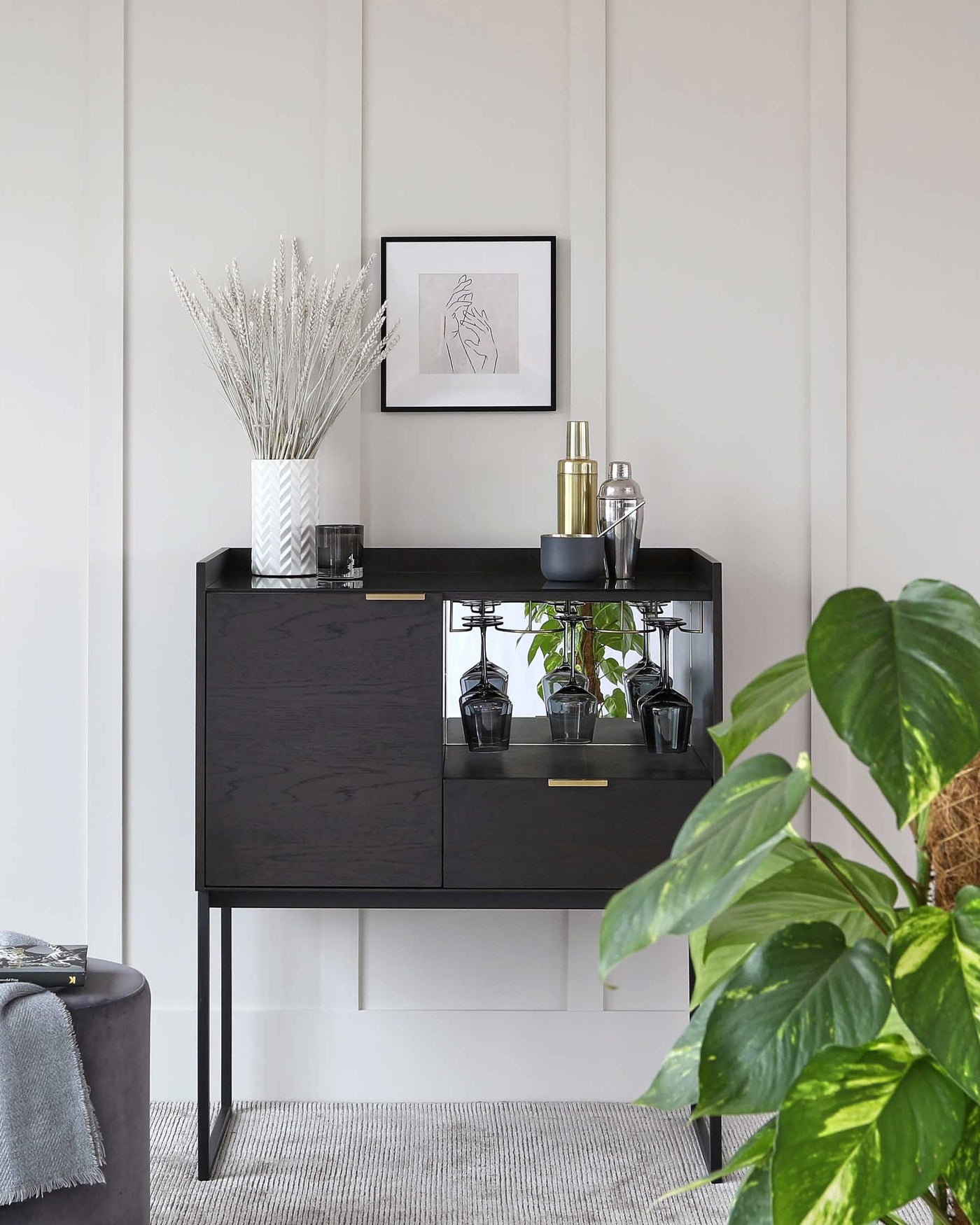 A modern black sideboard with brass handles and slender metal legs, featuring two cabinet doors and a shelf with glass panelling, displaying decorative items and houseplants.