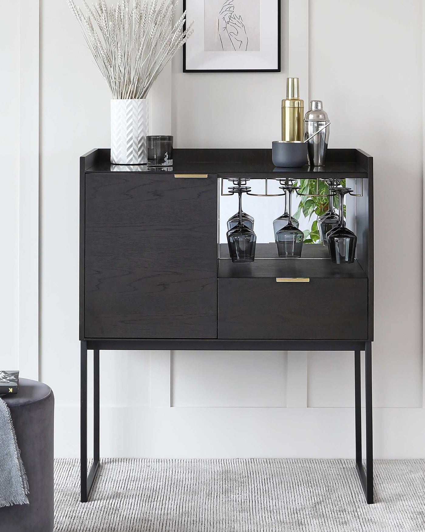 Modern black wooden sideboard with sleek metal handles and thin metal legs featuring two drawers and a lower shelf with glass holders, elegantly staged with decorative vases, cocktail shakers, and a framed artwork in the background.