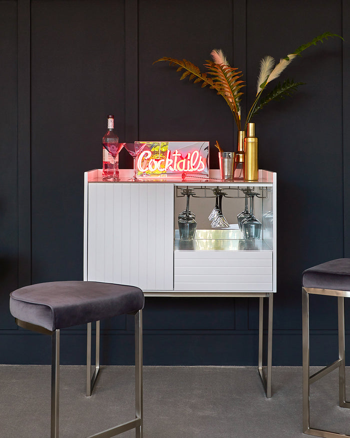 Modern home bar setup featuring a sleek white cabinet with a high-gloss finish and a minimalist design, complemented by two velvet-upholstered bar stools with slender metal legs, adding a touch of elegance and contemporary style to the space.