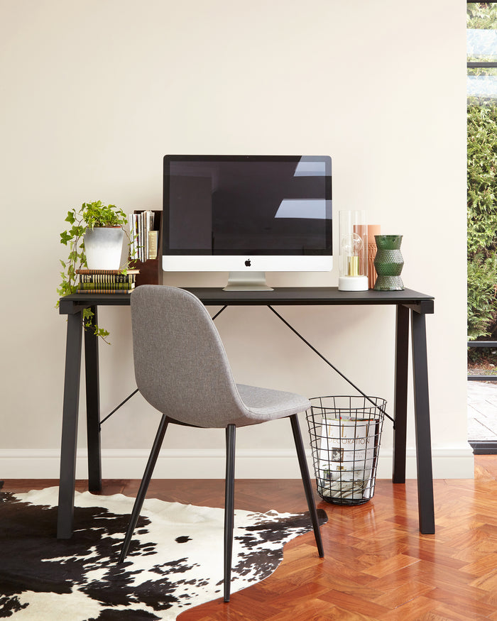 Modern minimalist home office setup featuring a sleek black desk with straight legs and a grey fabric upholstered chair with slender black metal legs. A wire trash can is positioned beside the desk.