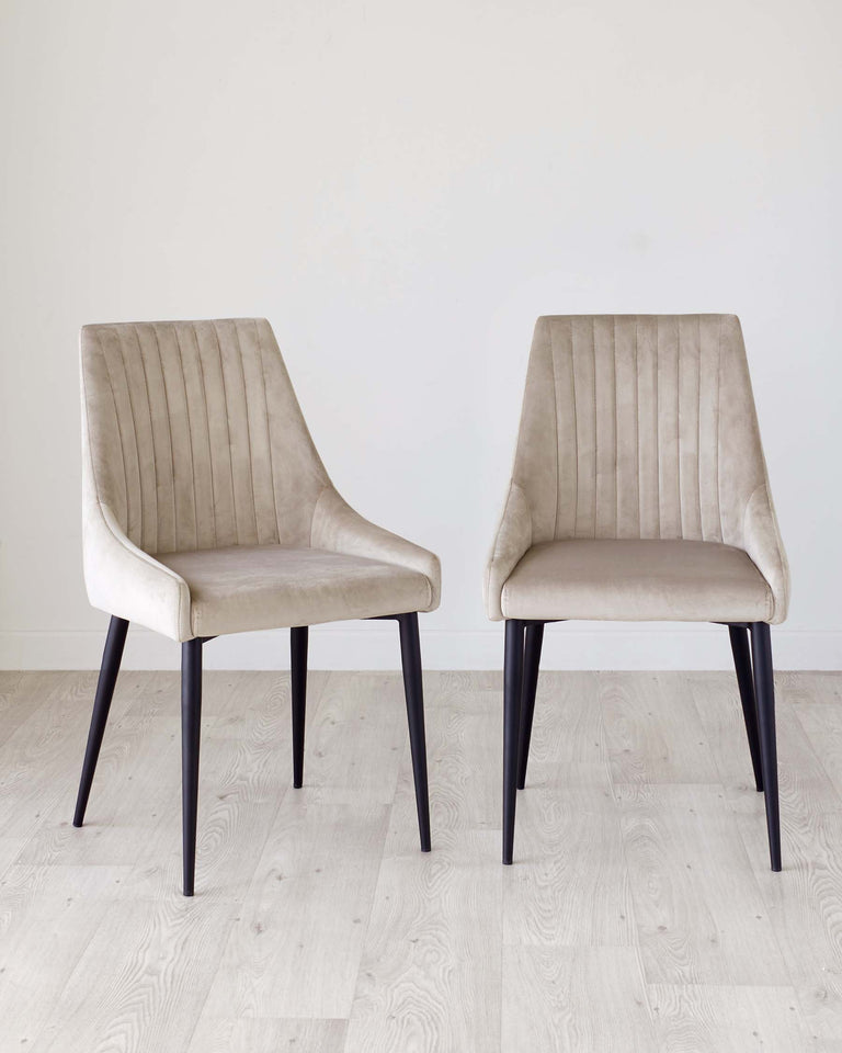 Two modern upholstered dining chairs with beige velvet fabric and vertical channel stitching on the backrest, featuring dark wooden legs, set on a light hardwood floor against a white wall.