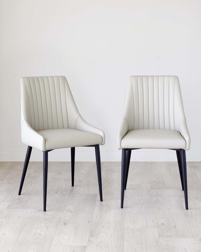 Two modern dining chairs with a sleek design, featuring cream-colored upholstered seats with vertical stitching on the backrest, and angled dark wooden legs. They are situated on a light hardwood floor against a plain white wall.