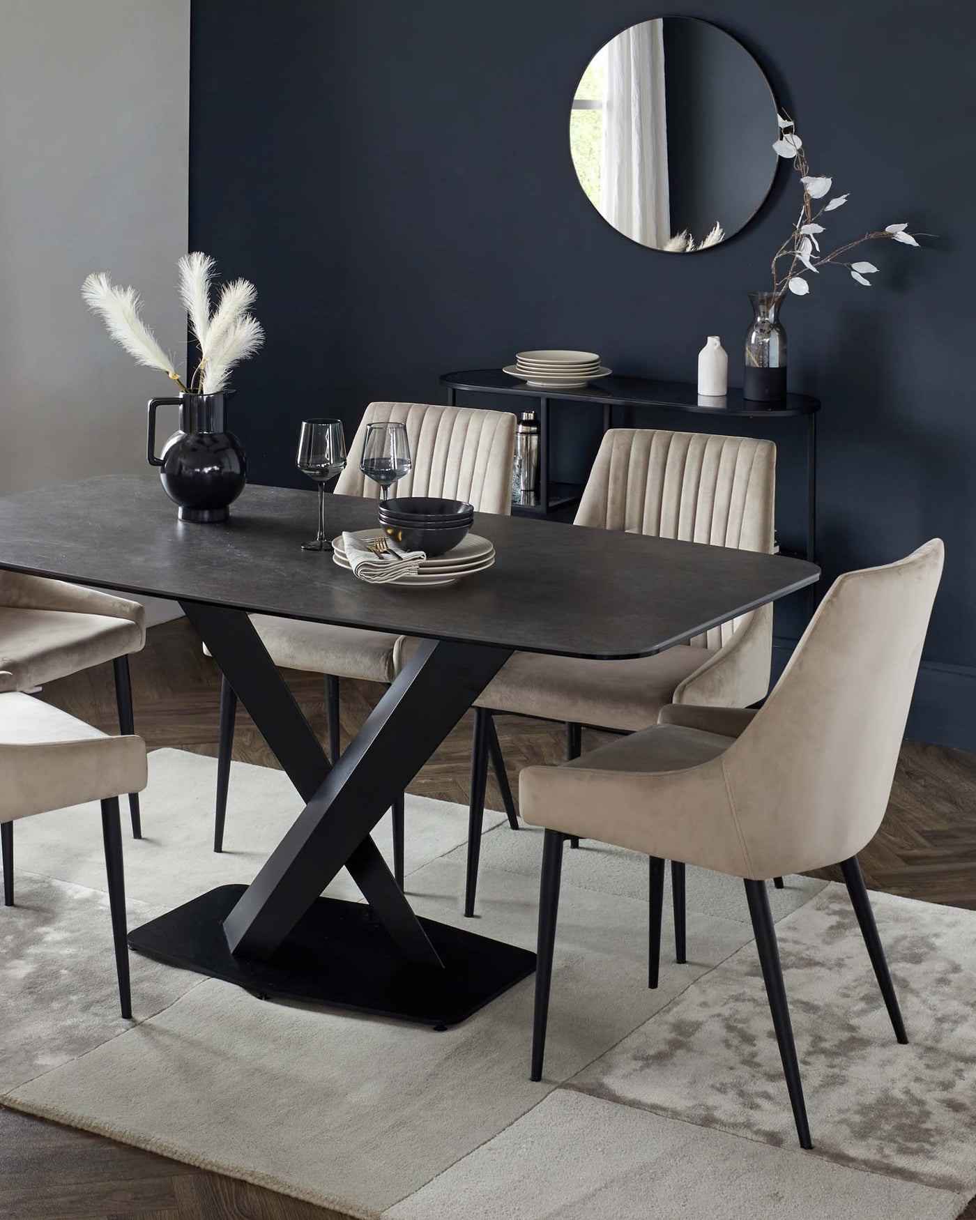 A contemporary dining room setup featuring a dark wood table with a unique X-shaped black metallic base, surrounded by four elegant taupe upholstered chairs with slender black legs. A minimal black console table with a circular mirror above it and decorative vases is visible in the background. The setting has a modern and sophisticated ambiance.