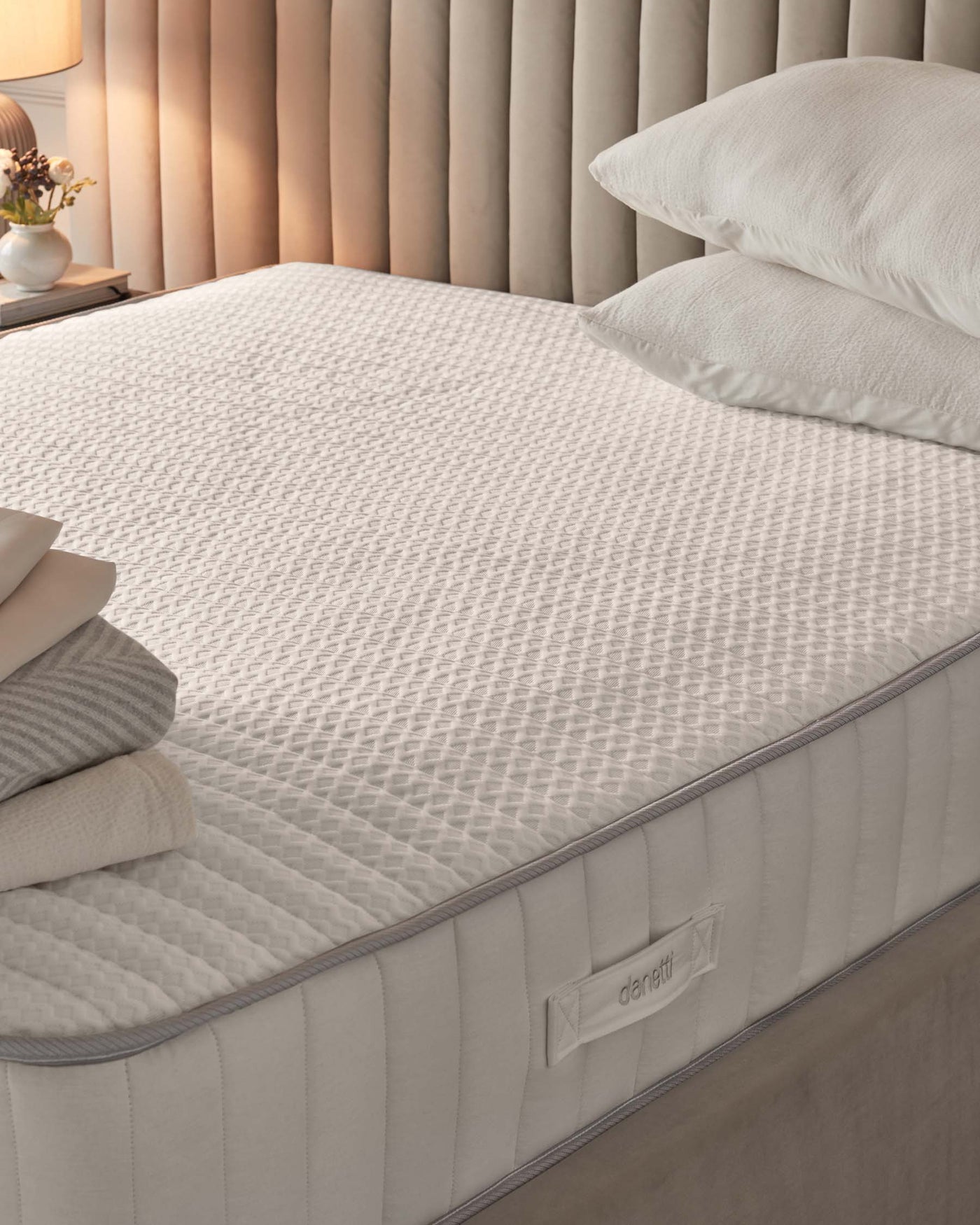 Luxurious queen-sized mattress with white quilted design and plush pillow-top. A pair of soft pillows with plain white covers are on top. Brand label visible on the mattress side. Elegant bedside with a neutral-toned lamp and flowers, set against a textured beige curtain backdrop.