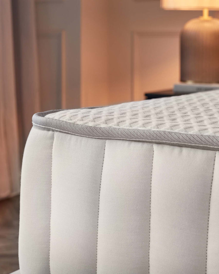 Close-up of a mattress featuring a white patterned top and white, vertically stitched sides, finished with grey piping.