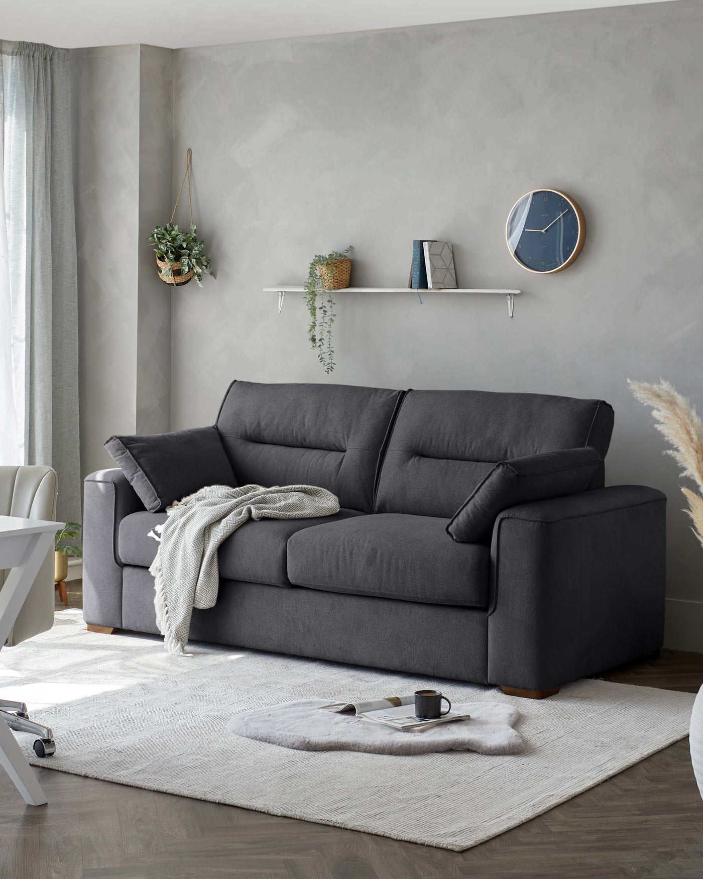 A contemporary charcoal grey fabric sofa with plush cushions and a clean-lined silhouette sits atop a light beige textured area rug, accented with a neutral-toned throw blanket casually draped on one side.