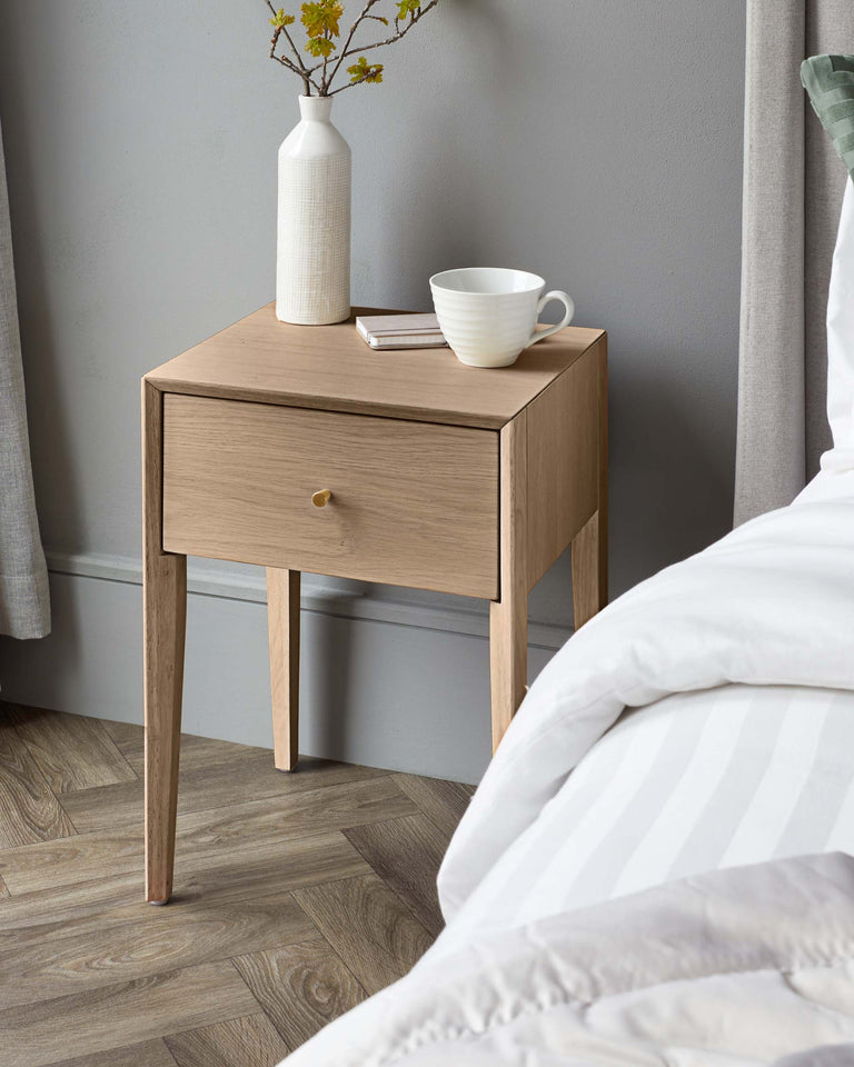 Modern light oak nightstand with a simplistic design featuring a smooth surface, a single drawer with a round, golden knob, and four straight, tapered legs. On the stand, there is a textured, white vase with yellow flowers, a stack of books, and a white ceramic cup.