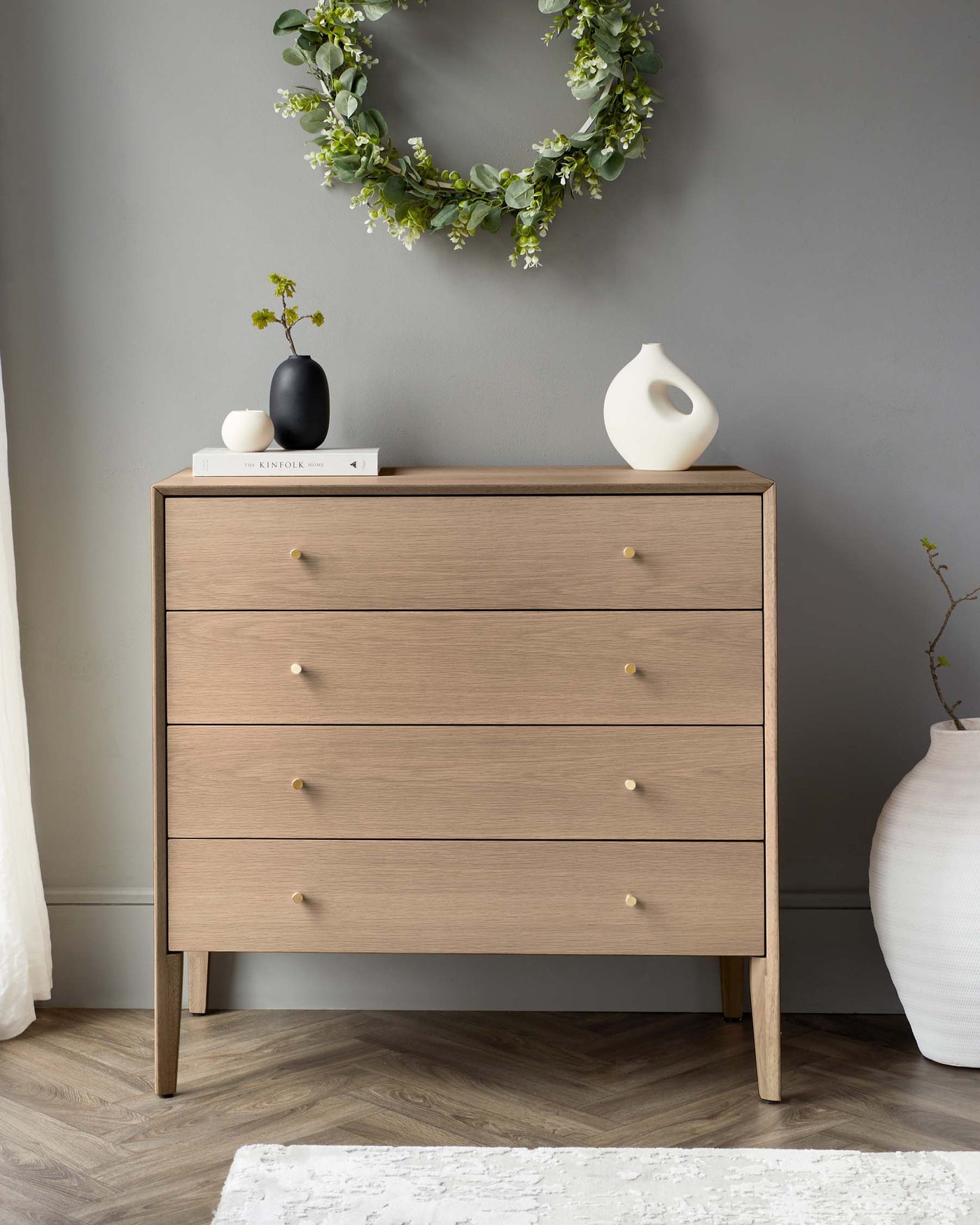 Modern four-drawer wooden dresser with round tapered legs and small round knobs, set against a grey wall with a green leafy wreath hanging above. A small white vase, black ceramic piece, and a book rest atop the dresser, next to a large white vase on the floor to the right.