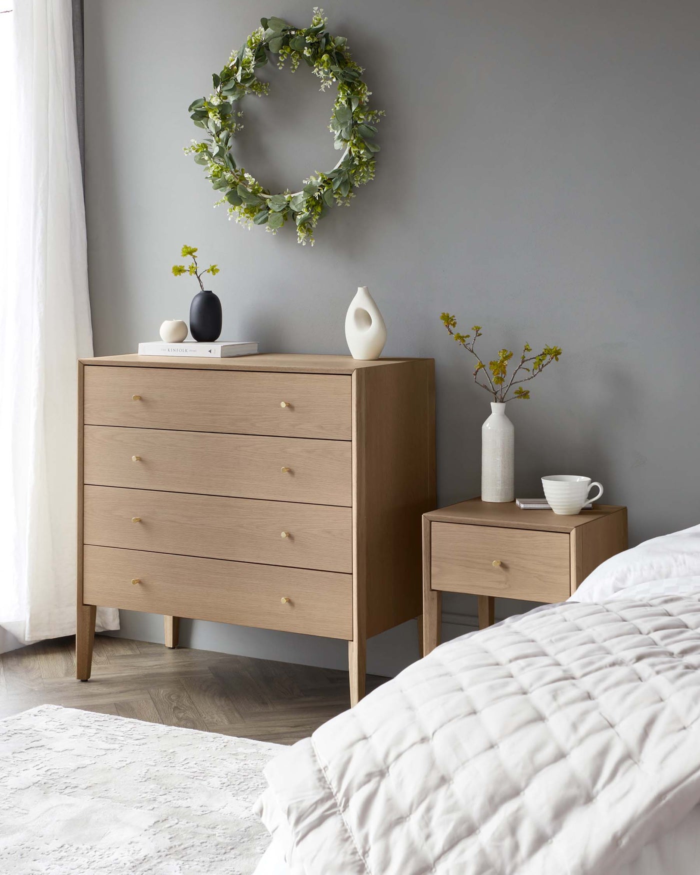 A modern wooden dresser with a four-drawer design featuring round knobs stands next to a matching single-drawer nightstand. Both pieces have a natural wood finish and tapered legs, creating a minimalist and contemporary look in the bedroom setting.