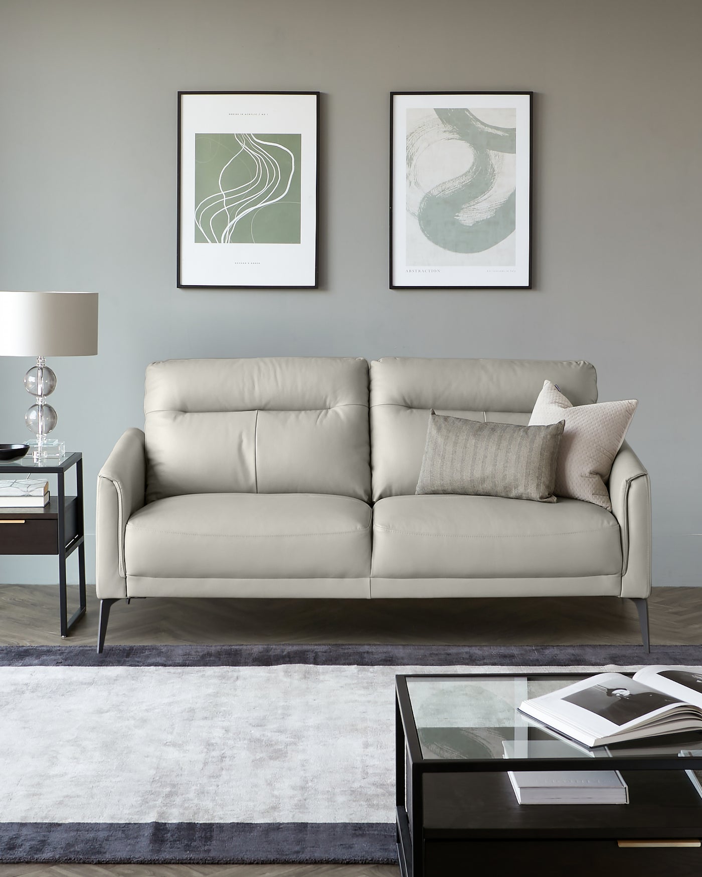 Contemporary light grey leather sofa with clean lines and metal legs, accompanied by a square black glass-top coffee table, standing on a grey and white area rug. A stylish table lamp with a translucent base is placed on a dark wood side table next to the sofa, and decorative grey-toned pillows accentuate the sofa's neutral palette.