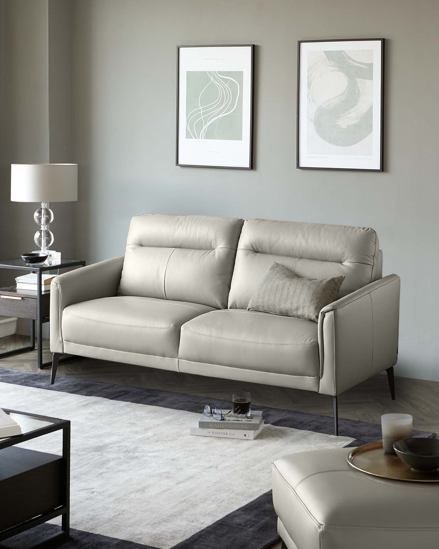 Modern light grey leather three-seater sofa with minimalistic design and clean lines, accompanied by a matching leather ottoman. A sleek side table with a glass top and dark frame stands adjacent to the sofa, supporting a chic table lamp. The neutral-toned area rug beneath complements the contemporary styling, framed by a dark wood coffee table with a reflective glass surface and minimal decor.