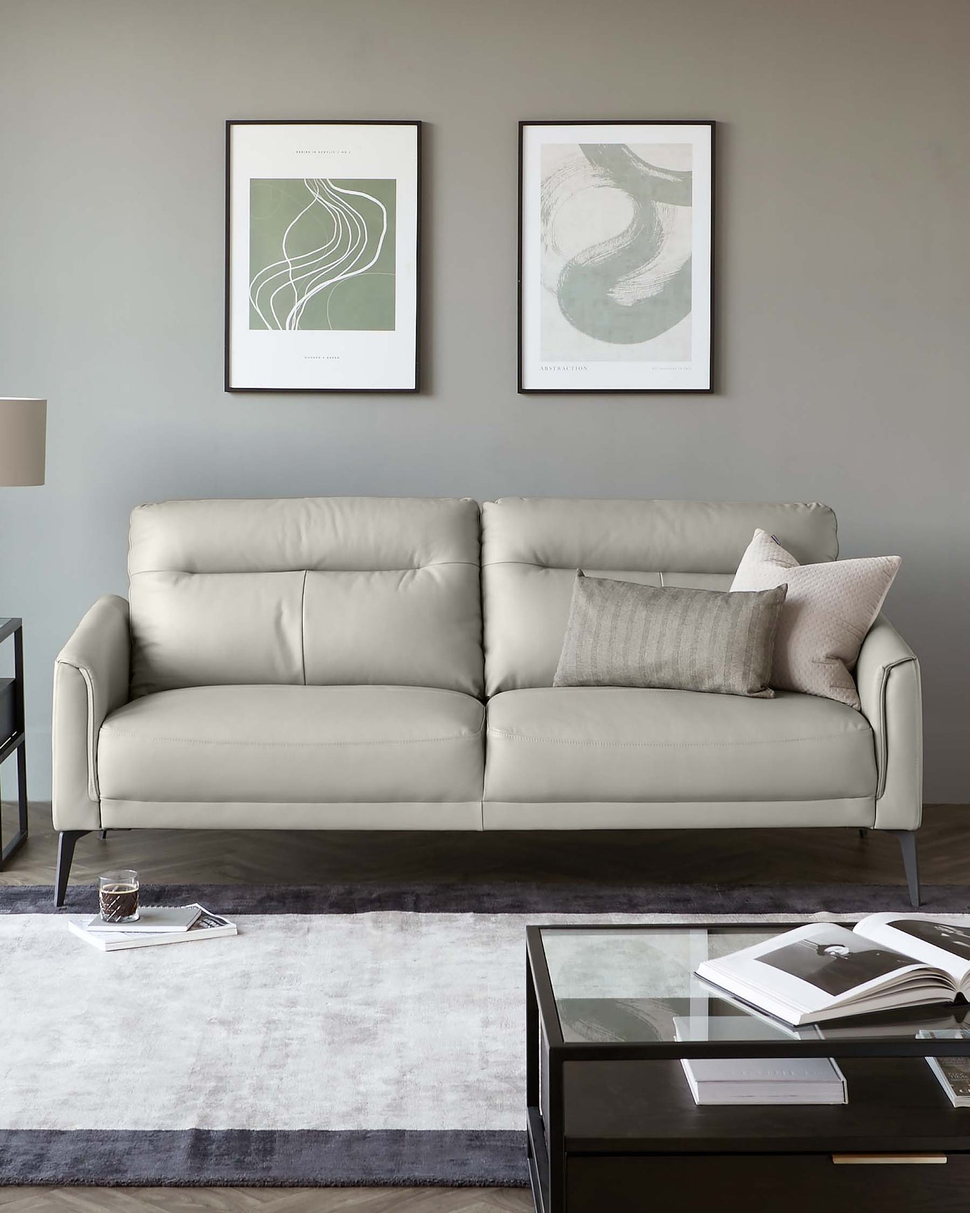 Elegant contemporary living room featuring a light grey upholstered sofa with plush cushions and sleek armrests, complemented by textured grey throw pillows. A dark wood and glass coffee table with a modern design sits on a large grey and lavender area rug, coordinating with the room's neutral colour palette.