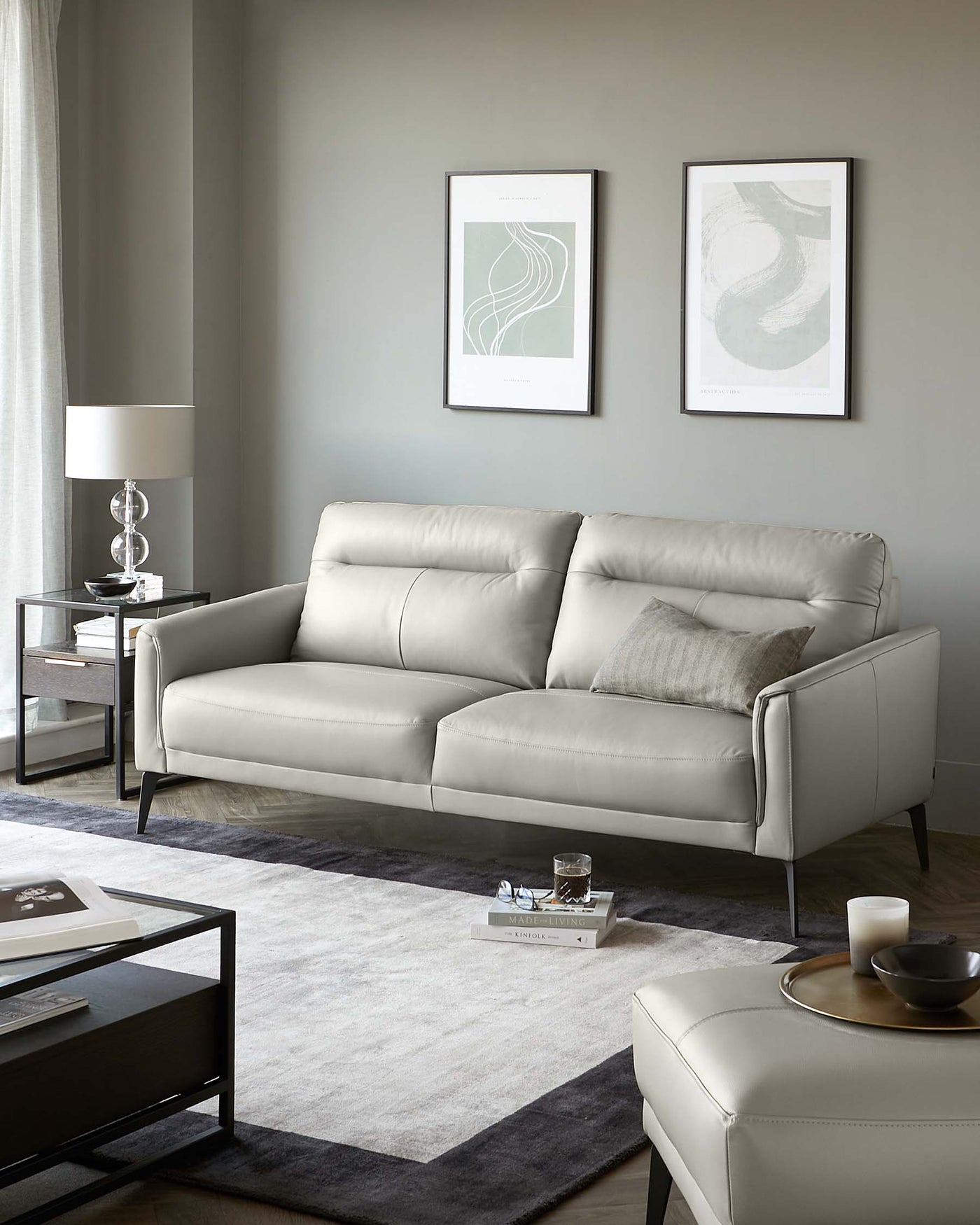 Elegant living room setup featuring a light grey leather sofa with clean lines and metallic legs, paired with plush cushions. Adjacent are two mirrored side tables with dark frames and a clear glass lamp on one of them. A dark wood coffee table with a metallic rim sits atop a two-tone grey area rug, completing the sophisticated space.
